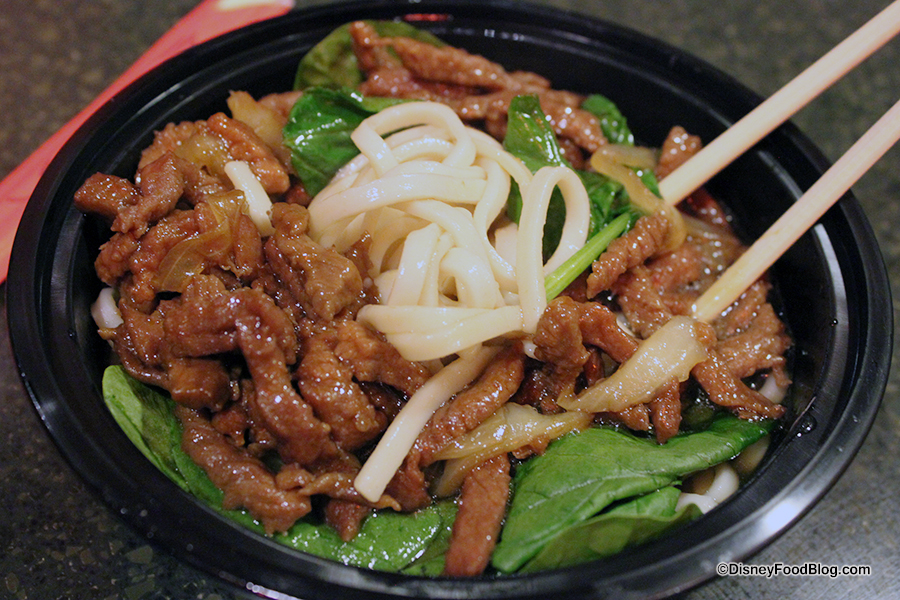 A close-up of a bowl filled with a noodle dish. The dish features sliced beef, sautéed onions, leafy greens, and thick noodles, all topped with chopsticks resting on the side. The ingredients are arranged in a black bowl against a dark background.