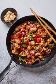 A black skillet filled with stir-fried chicken, red bell peppers, green onions, and peanuts, placed on a light textured surface. A pair of wooden chopsticks rest on the skillet's edge. A small bowl of peanuts sits beside the skillet.