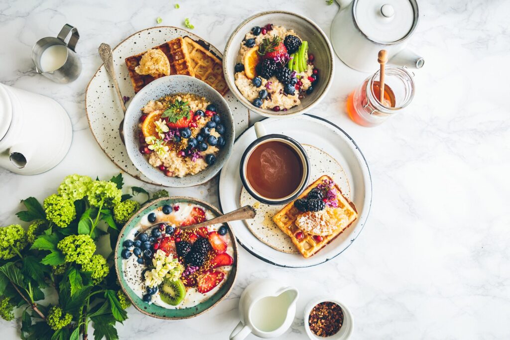 A top view of a breakfast spread, including vibrant bowls of oatmeal topped with berries, nuts, and sliced fruits, plates of waffles with cream and berries, a cup of coffee, a jar of honey, and a teapot. Fresh green flowers and leaves are also arranged on the white tabletop.