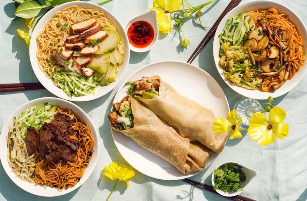 A table is set with colorful dishes including three bowls of noodles topped with meats and vegetables, a plate with two large spring rolls, a small dish of red sauce with a spoon, and some fresh herbs. Yellow flowers are scattered decoratively among the dishes.
